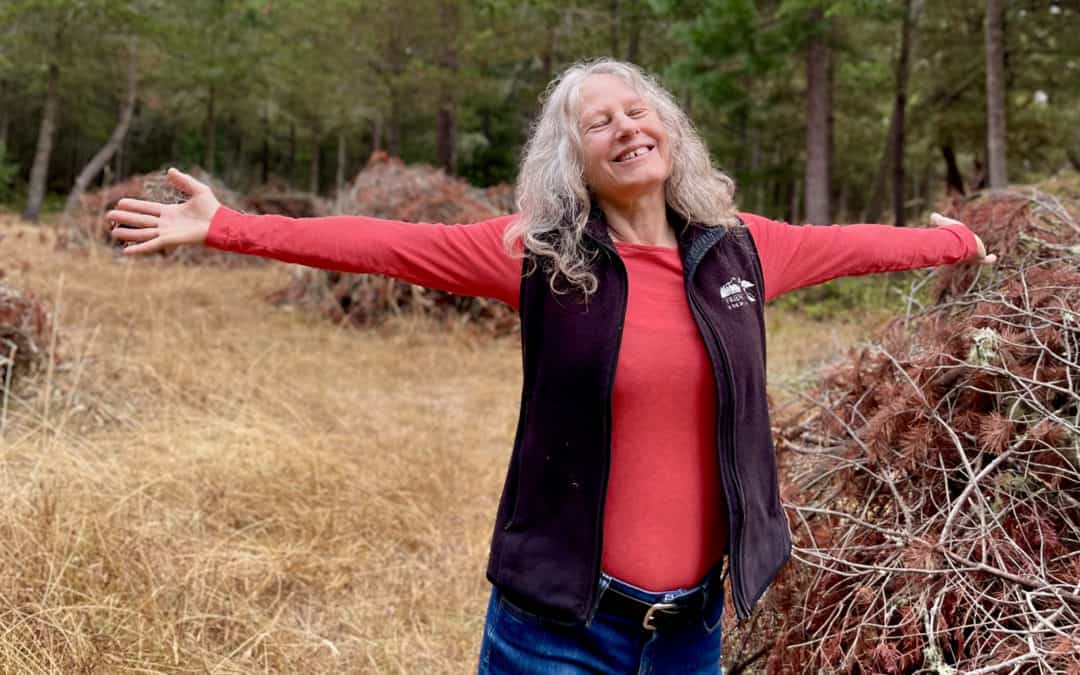 Older Woman standing in field surrounded by wood with arms extended wearing jeans, a long sleeve red shirt with dark vest