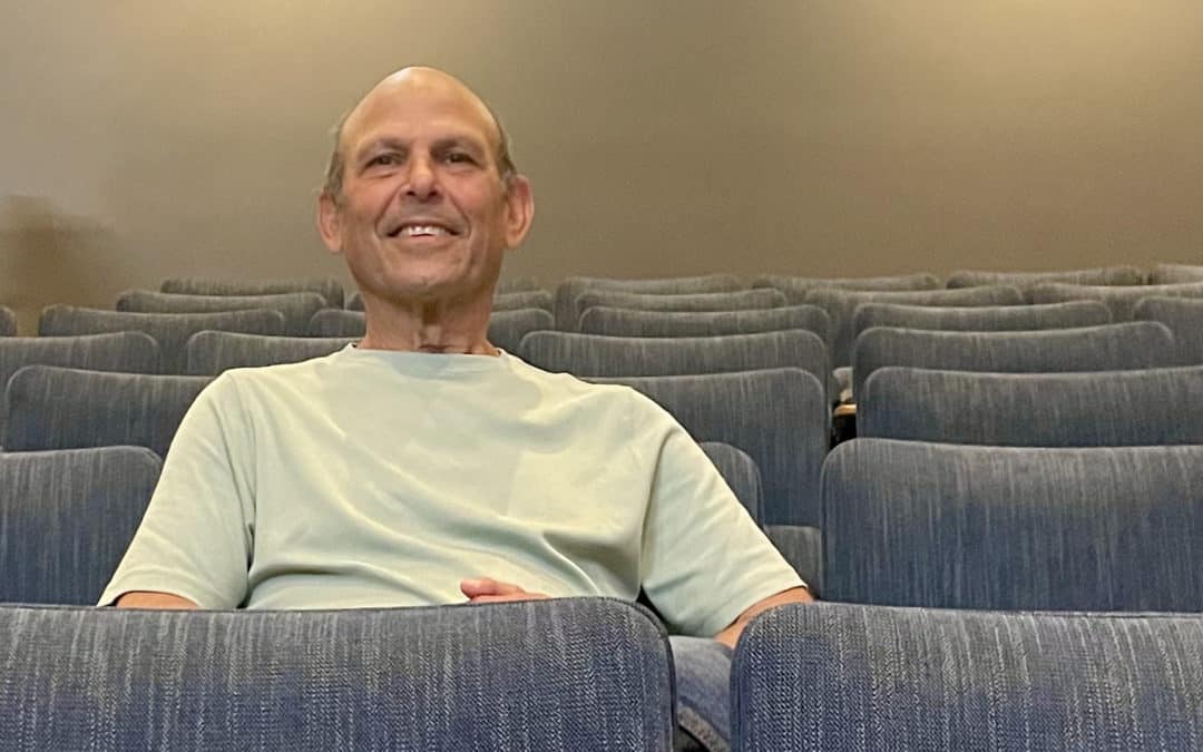 Older mostly bald headed main in t-shirt sitting in a row of theater chairs