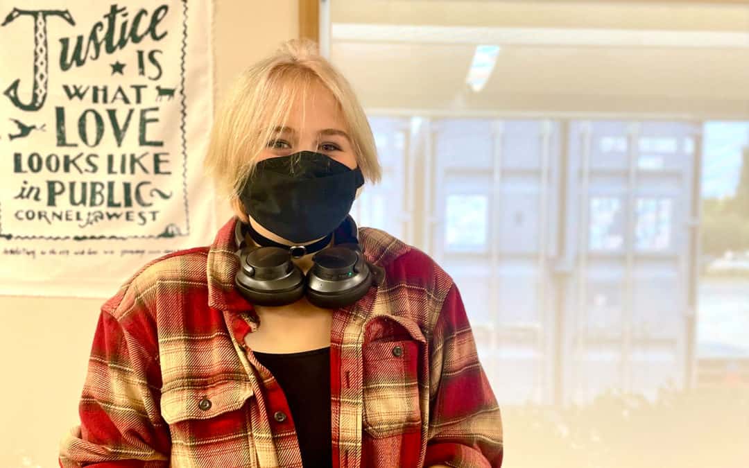 teen with short blonde hair wearing a red plaid shirt and black mask with headphones around her neck