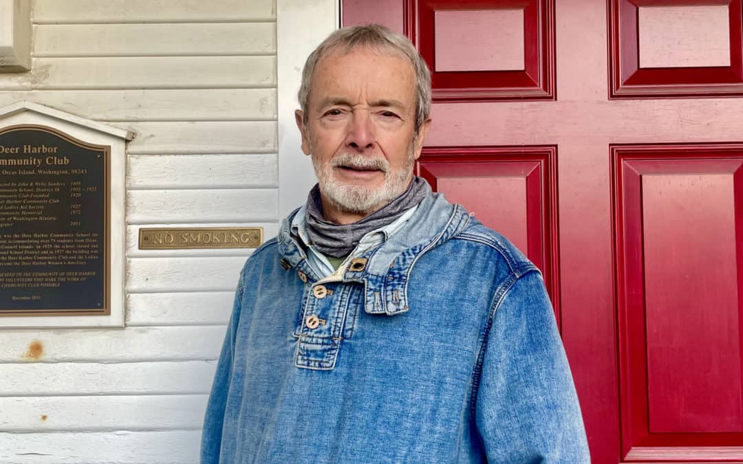 Older man with short gray hair and beard in jean shirt in front of red door