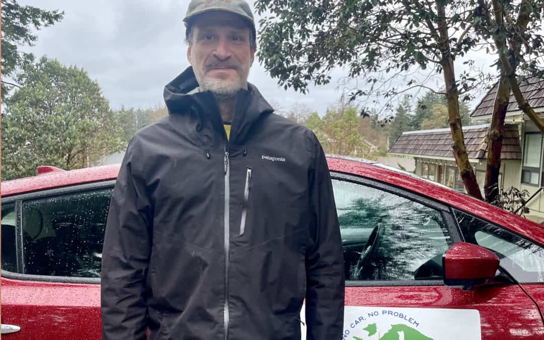 Middle aged/older man with graying beard with ball cap and rain jacket and car and gray skies a background