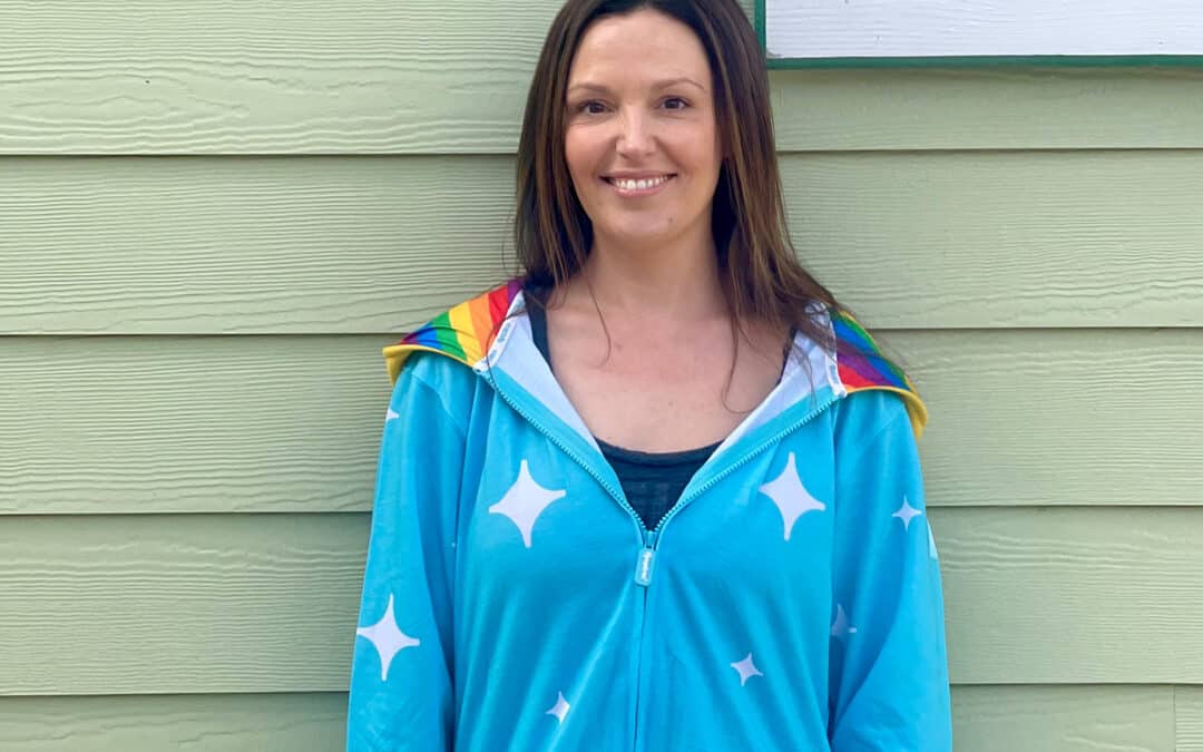 Smiling, middle-aged woman with long brown hair in a blue rainbow onesie with stars on it with a pale green siding in the background