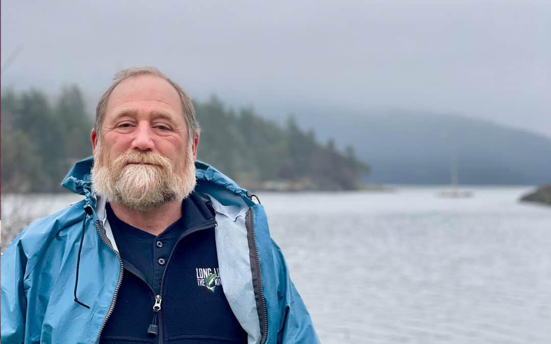 Older man in a blue jacket and shirt with gray hair and beard standing in front of the water with conifers blurred in the background